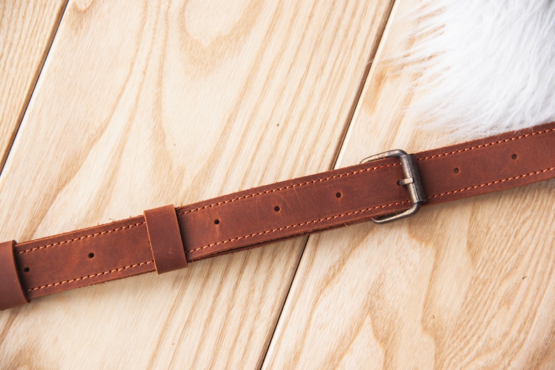 May include: A brown leather belt with a silver buckle. The belt is stitched with brown thread and has multiple holes for adjusting the size.