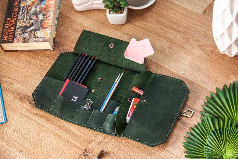 May include: A green leather pencil case with multiple pockets and compartments. The case is open and filled with pencils, pens, and other art supplies. The case has a metal clasp.