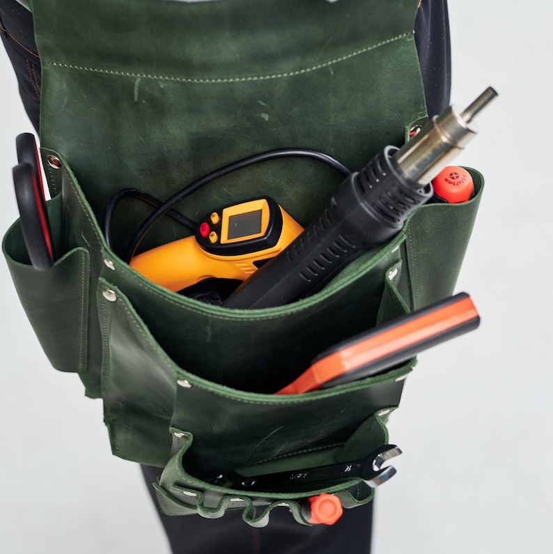 K&ouml;nnte beinhalten: Ein gr&uuml;ner Leder-Werkzeugg&uuml;rtel mit mehreren Taschen, der eine schwarze und gelbe Hei&szlig;luftpistole, ein rotes und oranges Werkzeug und einen schwarzen Schraubenschl&uuml;ssel enth&auml;lt.