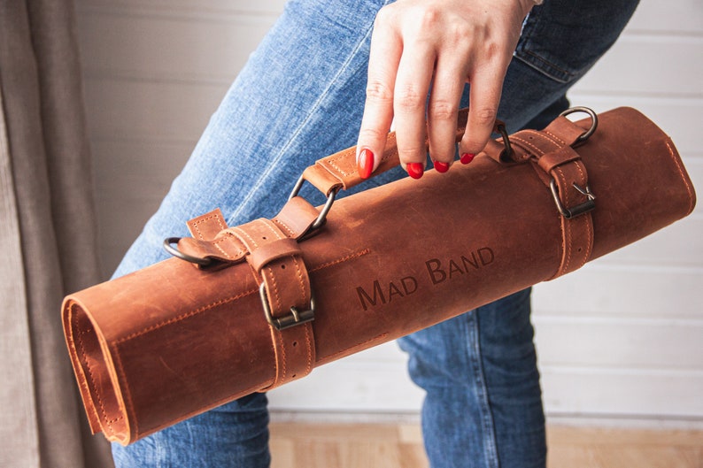 May include: A brown leather knife roll with a strap and buckle fastening. The roll has a handle and the text "MAD BAND" is embossed on the front.