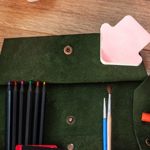 May include: A green leather pencil case with a flap closure and a snap button. The case is open and contains coloured pencils, paintbrushes, and glue sticks.