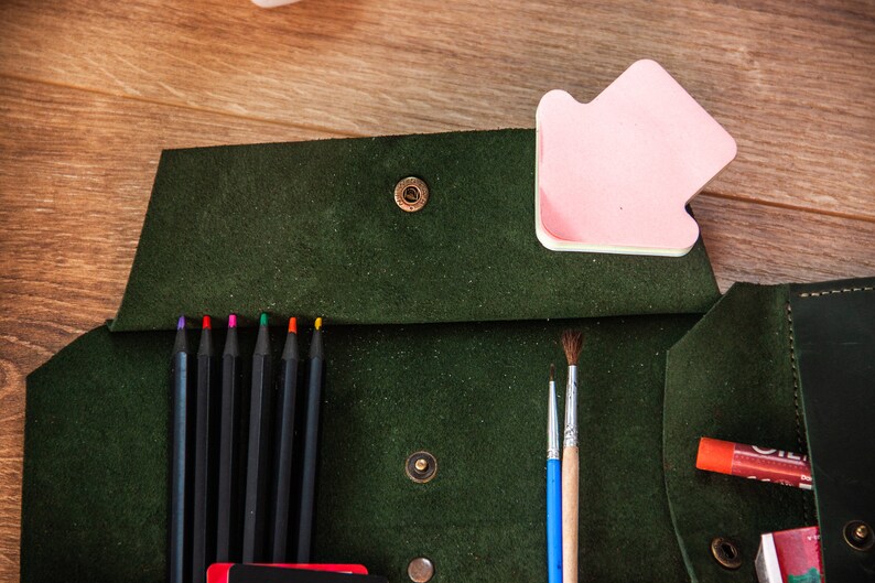 May include: A green leather pencil case with a flap closure and a snap button. The case is open and contains coloured pencils, paintbrushes, and glue sticks.