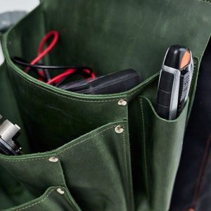 May include: A green leather tool bag with multiple pockets and a snap closure. The bag is filled with tools, including a black and silver utility knife and a black electrical tester.
