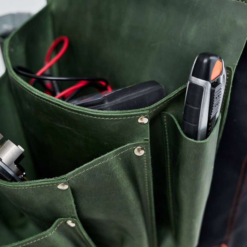 May include: A green leather tool bag with multiple pockets and a snap closure. The bag is filled with tools, including a black and silver utility knife and a black electrical tester.