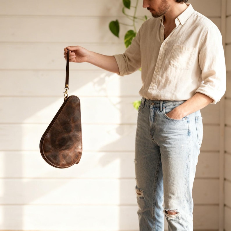 May include: A brown leather pouch with a handle, held by a person. The pouch has a unique, curved shape and a textured surface. The handle is a dark brown leather strap with a gold-colored clasp. The background is a white wall.