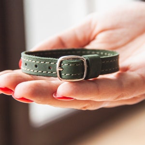 May include: A green leather bracelet with a silver buckle. The bracelet is stitched with white thread.