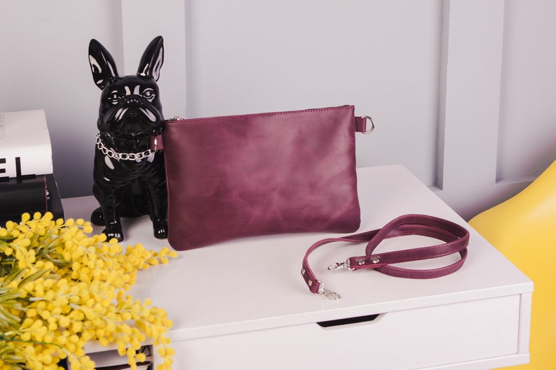 Peut inclure: Un sac &agrave; main en cuir bordeaux avec une bandouli&egrave;re en cha&icirc;ne argent&eacute;e. Le sac est pos&eacute; sur une surface blanche avec une figurine noire de bouledogue fran&ccedil;ais et un bouquet de mimosa jaune.
