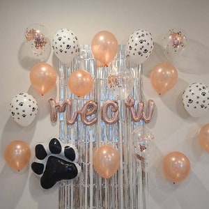 May include: A white wall decorated with balloons for a party. The balloons are in various colors, including rose gold, white, and clear. Some of the balloons have paw prints on them. There is a silver foil curtain behind the balloons. The word "meow" is spelled out in rose gold foil letters. There is a black paw print balloon in the lower left corner.