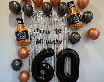 May include: A silver and black birthday party decoration with black balloons shaped like the numbers 60, black and gold balloons shaped like bottles, and a silver tinsel backdrop with the words "cheers to 60 years".