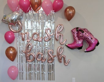 May include: A silver and white streamer backdrop with the words "nash bash" in rose gold foil letters. There are pink and rose gold balloons, and a pair of pink cowboy boot balloons.