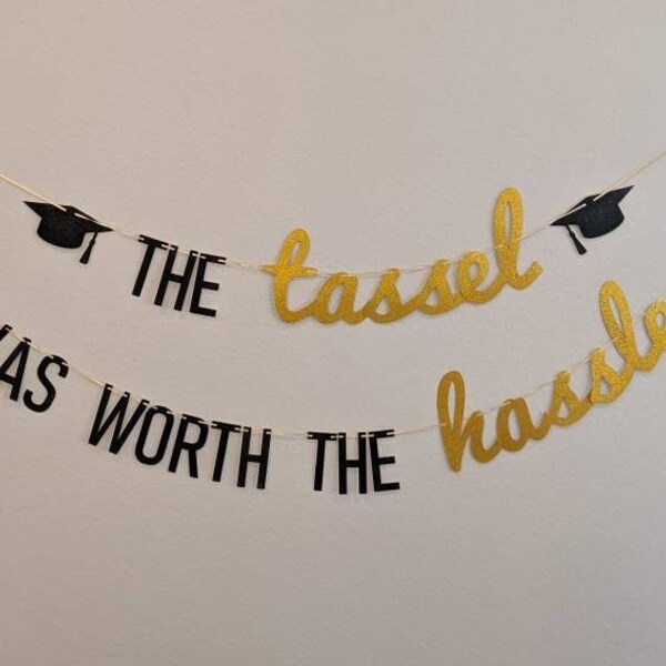 May include: A graduation banner with the text "THE tassel WAS WORTH THE hassle" in black and gold lettering. The banner includes black graduation cap cutouts and is strung on a thin white string.
