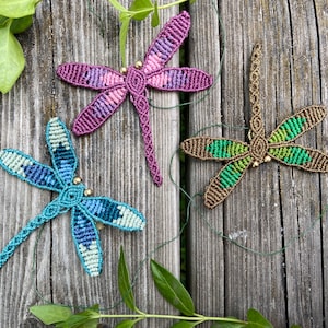 Pode incluir: Três tapeçarias de libélula de macramé em diferentes cores. A primeira libélula é roxa e rosa, a segunda é azul e verde, e a terceira é marrom e verde.