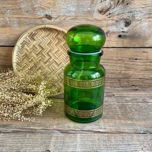 Peut inclure: Un bocal d'apothicaire en verre vert vif avec un couvercle assorti. Le bocal présente une bande décorative dorée au milieu. Un plateau en bambou tressé et des fleurs séchées sont en arrière-plan. Le bocal est sur une surface en bois.