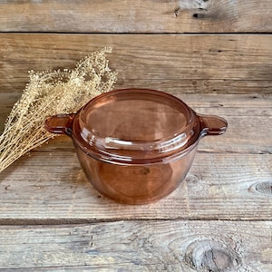 Può includere: Una pirofila in vetro marrone con coperchio abbinato. La pirofila ha due manici ed è appoggiata su una superficie di legno. La pirofila è di un caldo colore marrone e sembra essere realizzata in vetro resistente.