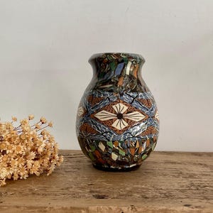 Może przedstawiać: Pękata ceramiczna waza z wielokolorowym wzorem mozaiki składającym się z geometrycznych kształtów i abstrakcyjnych linii. Wzór zawiera centralny motyw rombu z ciemnym środkiem, otoczony niebieskimi, brązowymi, zielonymi i czarnymi segmentami.