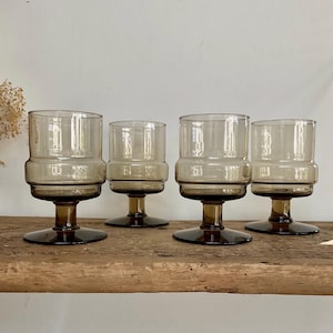 Op de afbeelding: Een set van vier bijpassende drinkglazen gemaakt van doorschijnend rookkleurig glas. Elk glas heeft een brede, cilindrische kom met een duidelijke horizontale rand in het midden, ondersteund door een korte, dikke steel en een brede ronde voet.