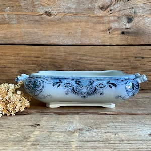 Peut inclure: Un plat de service rectangulaire en céramique avec une base blanche ornée de motifs floraux et de volutes bleus complexes. Le récipient comprend deux petites anses sur les côtés et repose sur une base surélevée au design décoratif.