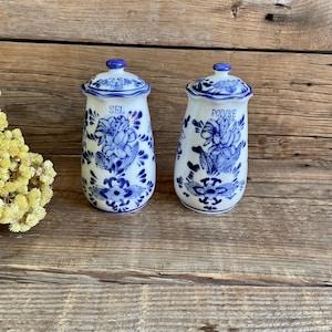 Op de afbeelding: Twee witte keramische zout- en peperstrooiers met blauwe bloemmotieven. De strooiers hebben een cilindrische vorm met een deksel en zijn gemarkeerd met "SEL" en "POIVRE". Ze staan op een houten oppervlak.
