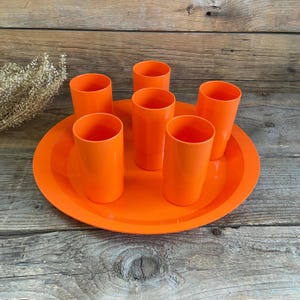 Könnte beinhalten: Ein Set aus sechs orangefarbenen Trinkgläsern aus Kunststoff auf einem passenden runden Tablett. Die Gläser sind zylindrisch und gleichmäßig auf dem Tablett angeordnet. Das Tablett und die Gläser sind in einem leuchtenden, einfarbigen Orange gehalten.