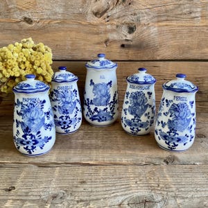 Puede incluir: Cinco botes de especias de cerámica blanca con diseños florales azules y tapas. Cada bote está etiquetado con un nombre de especia diferente: Paprika, Cannelle, Ail, Kok-Muscade y Persil. Los botes están dispuestos sobre una superficie de madera.