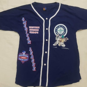 May include: A navy blue baseball jersey with white piping. The jersey has the words "American League" vertically down the left side, "Western Division Champs" on the front, and a Seattle Mariners logo with a cartoon character on the right side. The jersey also has a patch that says "Division Series 1995".