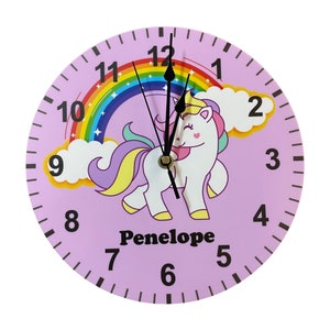 May include: A pink and purple unicorn clock with a rainbow and clouds. The clock face has black numbers and hands. The clock is personalised with the name "Penelope".