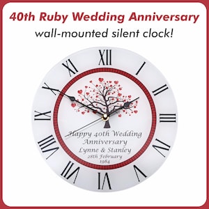 May include: A round, wall-mounted silent clock with a white face, black Roman numerals, and a red border. The clock has a tree design with heart-shaped leaves and the text "Happy 40th Wedding Anniversary Lynne & Stanley 28th February 1984".