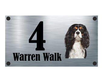 May include: A silver metal house number sign with the number 4 and the text "Warren Walk" in black. A black, white, and brown Cavalier King Charles Spaniel dog is pictured on the right side of the sign.
