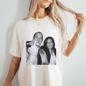 Puede incluir: Una camiseta blanca con una foto en blanco y negro de dos personas. La persona de la izquierda está haciendo el dedo medio.