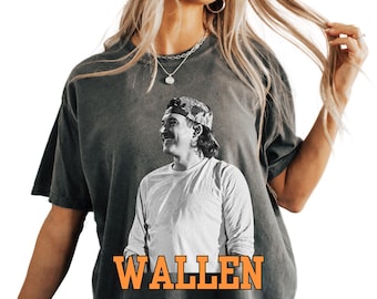 May include: Dark grey t-shirt featuring a black and white image of a man wearing a camouflage hat and long-sleeved shirt. The word "WALLEN" is printed in orange block letters across the bottom of the image.