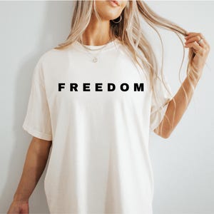 May include: Cream-colored short-sleeved t-shirt with the word "FREEDOM" printed in bold, black capital letters. The shirt is made of a soft material and has a relaxed fit. The overall style is casual and comfortable.