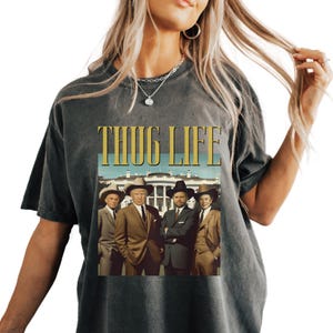 Puede incluir: Una camiseta gris oscura con un estampado gráfico vintage de cuatro hombres con sombreros de vaquero frente a la Casa Blanca. El texto "THUG LIFE" está impreso en dorado sobre la imagen.