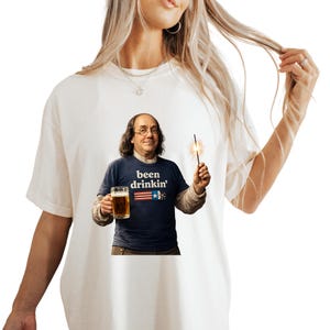 May include: Cream-colored t-shirt featuring a graphic of Benjamin Franklin holding a beer and a sparkler. The navy blue shirt he's wearing says "been drinkin'" with an American flag design. A fun and patriotic design.