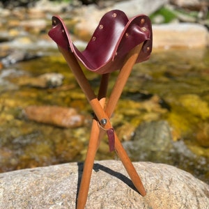 Puede incluir: Un taburete de camping de cuero burdeos con una base de trípode de madera. El taburete está sentado en una roca grande.