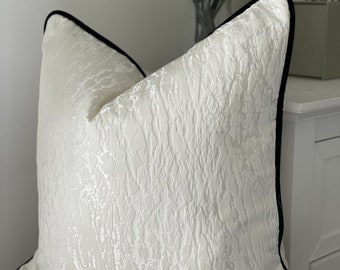 May include: A white throw pillow with a textured surface and a black trim. The pillow is sitting on a white surface.