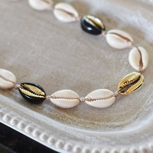 Könnte beinhalten: Eine Halskette aus weißen und schwarzen Muscheln mit goldenen Akzenten. Die Kette ist auf einer braunen Schnur aufgefädelt.