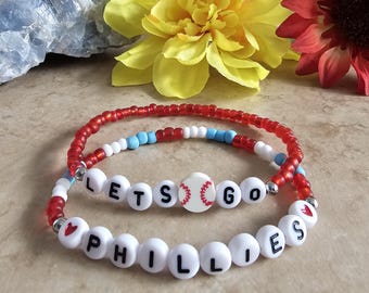 May include: Two beaded bracelets. One bracelet has red, white, and blue beads. The other bracelet has white beads with the words "LETS GO PHILLIES" spelled out. A baseball bead is in the center of the top bracelet.