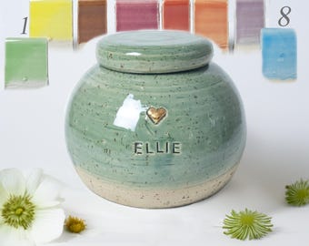 May include: A handcrafted, round, green ceramic urn with a lid, featuring a gold heart and the name "ELLIE." The urn is speckled with a light beige base. Eight color samples are displayed above the urn.