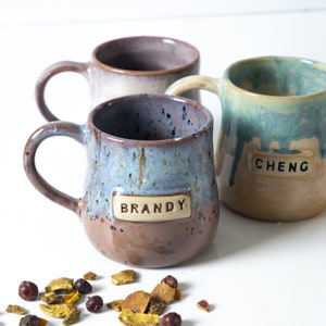 Può includere: Tre tazze in ceramica con motivi di smalto unici e nomi personalizzati. Le tazze sono Brandy, Cheng e una terza tazza con una smaltatura viola chiaro.