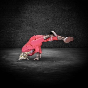 May include: A cat with a human body is breakdancing. The cat is wearing a red jumpsuit and black and white sneakers. The cat is balancing on its head and hands, with its legs extended in the air. The background is a dark gray wall and floor.