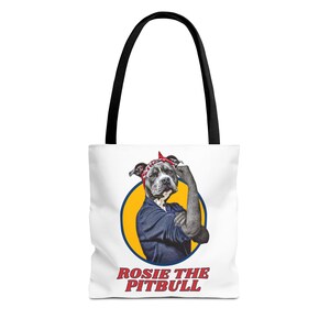 Puede incluir: Bolso tote blanco con asas negras. Presenta un gráfico de un pitbull musculoso, con un pañuelo rojo y una camisa azul. El perro está dentro de un círculo amarillo con el texto "ROSIE THE PITBULL" en rojo.