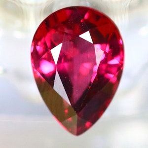 Puede incluir: Una gema facetada en forma de pera, de un intenso color rojo. El corte de la gema revela múltiples facetas que reflejan la luz, creando un brillo brillante. La gema se encuentra sobre un fondo borroso y claro.