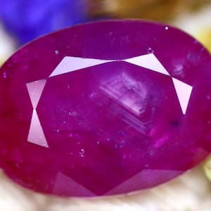 May include: A faceted, oval-shaped ruby gemstone. The gem is a deep, saturated red-pink color with internal reflections. The cut features multiple facets that reflect light, creating a brilliant appearance. The gemstone is set against a blurred background.