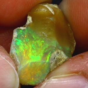 Op de afbeelding: Een ruwe, groene en oranje opaal edelsteen met een natuurlijke, onregelmatige vorm. De opaal wordt in een hand gehouden.