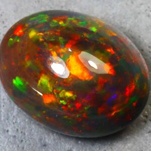 K&ouml;nnte beinhalten: Ein schwarzer Opal-Edelstein mit einem Cabochon-Schliff. Der Opal zeigt eine Vielzahl von Farben, darunter Rot, Orange, Gr&uuml;n und Lila.
