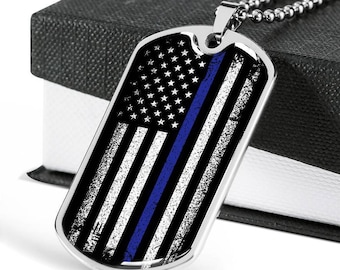 May include: A silver dog tag necklace featuring a distressed American flag design with a thin blue line running vertically down the centre. The necklace is on a silver chain and is displayed in a black box.