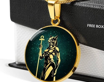 May include: Gold necklace with a round pendant featuring a black silhouette of a woman with a cat head holding a staff with an ankh symbol on top. The pendant has a green background with gold glitter.