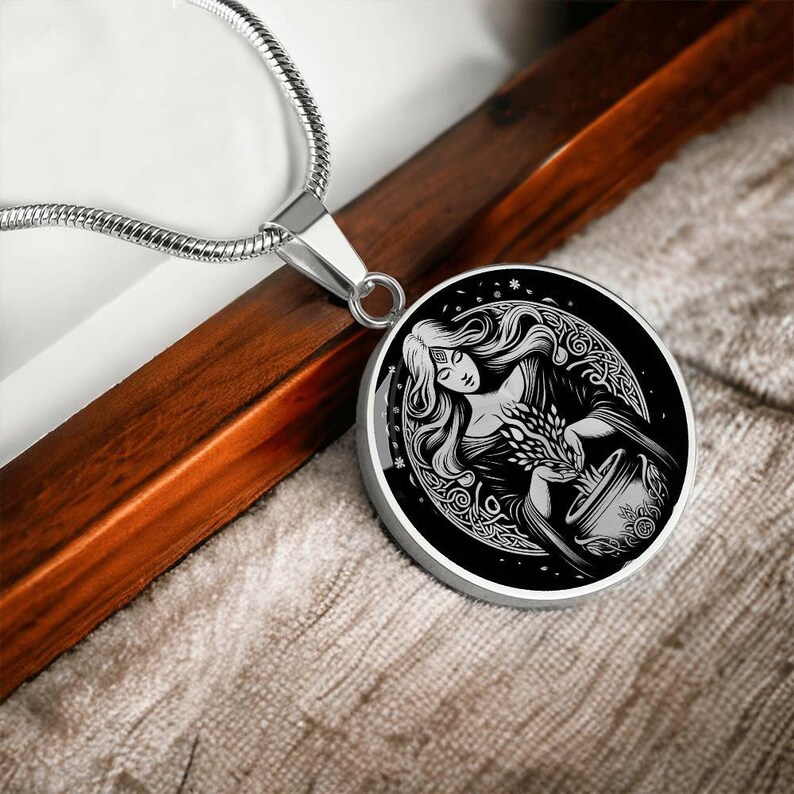 May include: A silver necklace with a round pendant featuring a black and white illustration of a woman holding a cauldron. The woman is surrounded by a Celtic knot design.