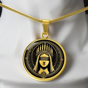 May include: Gold necklace with a round pendant featuring a black and gold illustration of a woman with a blindfold and a crown of candles. The pendant has a detailed circular design.