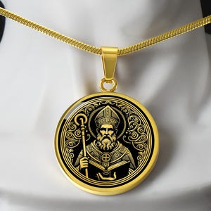 Op de afbeelding: Een gouden ketting met een ronde hanger met een zwart-gouden illustratie van een heilige met een baard en een miter. De heilige houdt een boek en een staf vast.